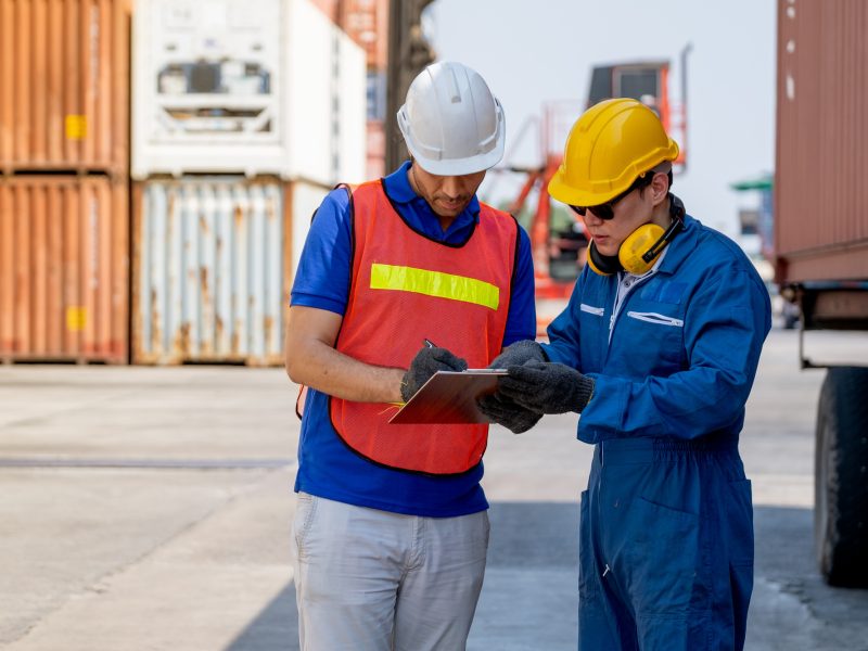 Cargo container worker and engineer discuss together with data in document on clipboard